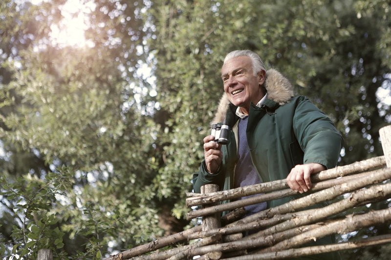 A mature man is bird-watching with binoculars in a park. This is a fantastic activity for promoting wellbeing in dementia