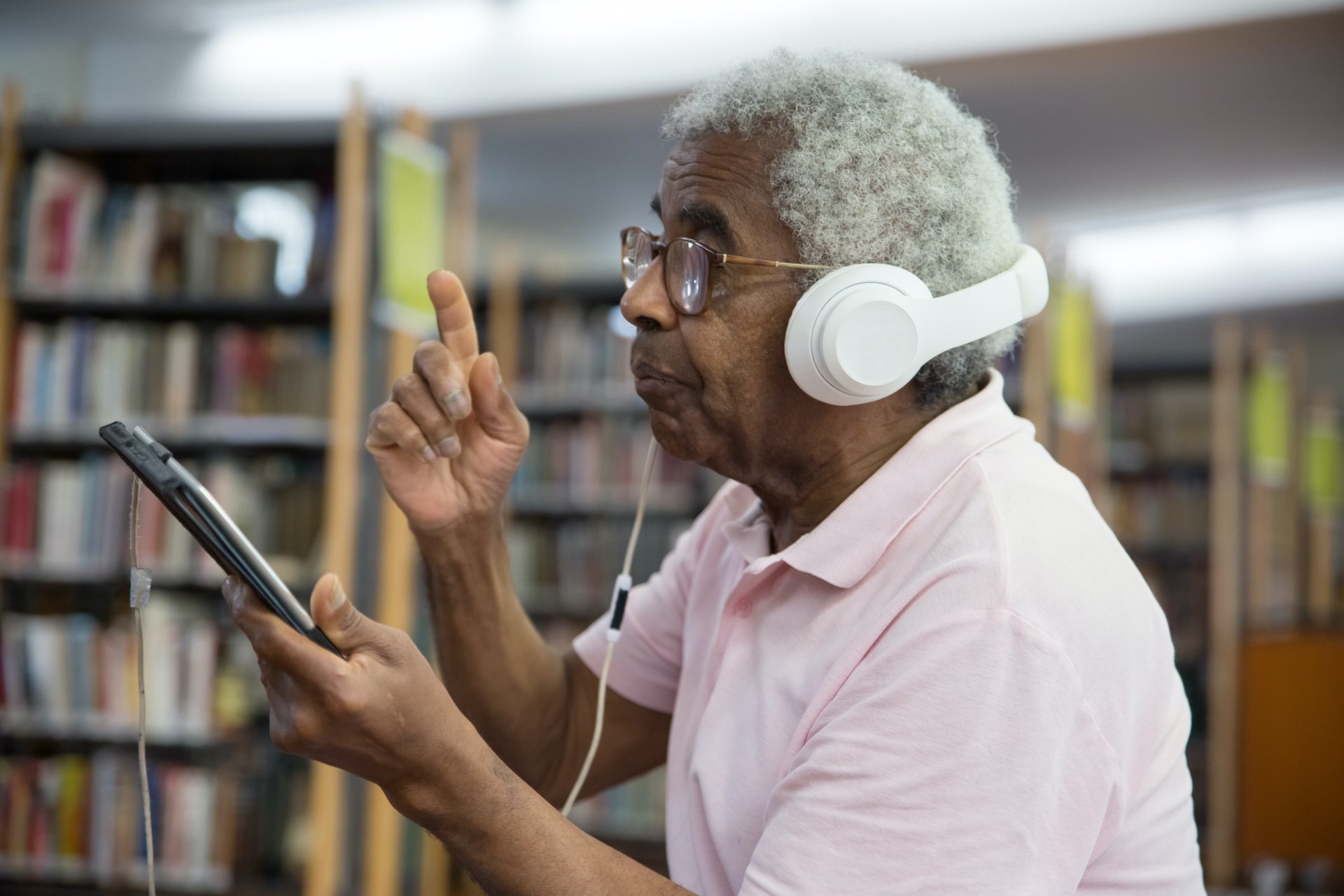 An elderly man, wearing a pale pink polo shirt, listening to music on his phone through white headphones in a library