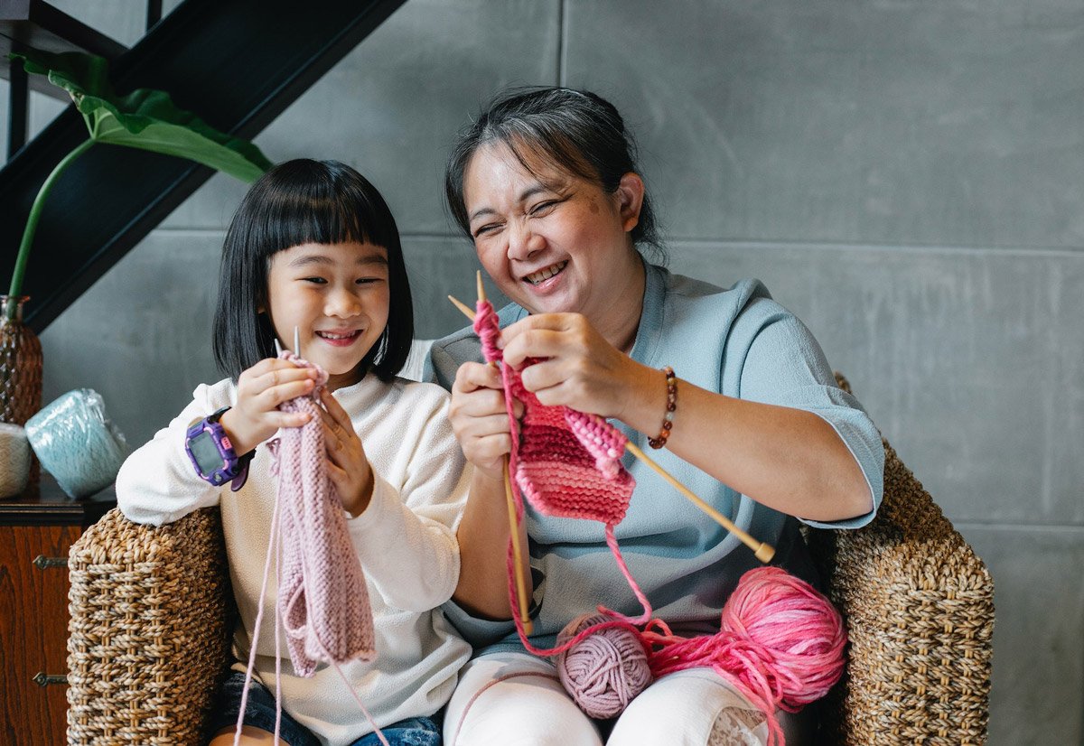 A granddaughter with her granddaughter are sitting on a sofa. The grandmother is showing her granddaughter how to knit. This is a dementia-friendly activity.